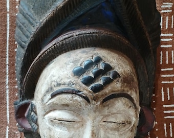 Pode incluir: Uma máscara de madeira com um rosto esculpido, olhos fechados e um sorriso largo. A máscara é pintada em tons de marrom, preto e branco. A máscara tem um cocar decorativo com um padrão geométrico.