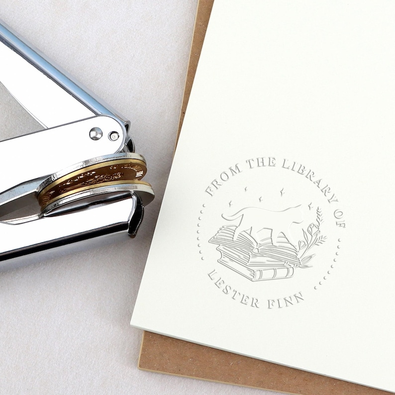 May include: A silver and gold metal embosser next to a cream-colored card with a circular design. The design includes a dog on a stack of books, the text "FROM THE LIBRARY OF LESTER FINN".