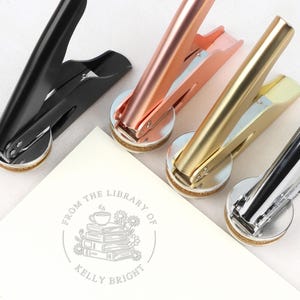 May include: Close-up of four metal library seal stamps in black, rose gold, gold, and silver. A white card with a circular design that reads "From the Library of Kelly Bright" is in the foreground. The design includes books, flowers, and a teacup.