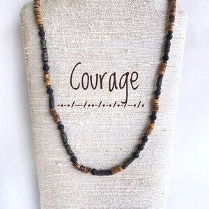 Könnte beinhalten: Eine braune und grüne Perlenkette mit dem Wort "Courage" auf weißem Hintergrund.