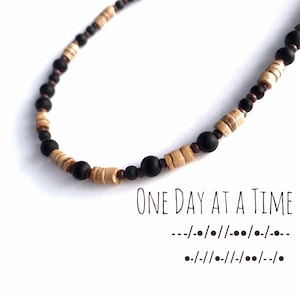 Op de afbeelding: Een kralenketting met zwarte en bruine houten kralen. Onder de ketting staat de tekst "ONE DAY AT A TIME" en twee regels morsecode.