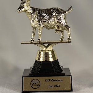 Könnte beinhalten: Goldene Trophäe mit einer nach links gerichteten Ziegenfigur auf einem schwarzen Sockel. Der Sockel hat eine goldene Plakette mit dem Text "DCF Creations Est. 2024".