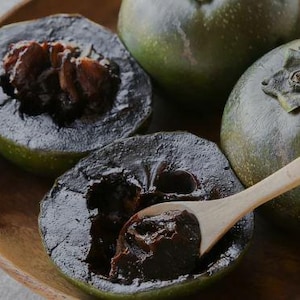 May include: Close-up of three dark green fruits with a brown, almost black, flesh inside. The fruit is cut open and the flesh is visible. A wooden spoon is scooping out the flesh of one of the fruits.