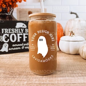 May include: A brown glass jar with a bamboo lid and a clear straw. The jar has a white ghost design with the text "Your Design Here" and "Estd 2024" on it. The jar is sitting on a wooden table with a black sign that says "Freshly Brewed Coffee" and a white pumpkin in the background.
