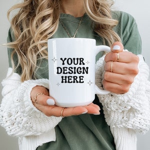 May include: White ceramic mug with a black design that says "YOUR DESIGN HERE" with stars on either side of the text. The mug is being held by a person wearing a white cardigan and a green shirt.