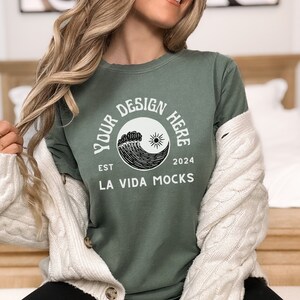 May include: Sage green t-shirt with a round graphic design featuring a wave, sun, and the text "YOUR DESIGN HERE" and "LA VIDA MOCKS". The shirt is worn with a cream-coloured cable knit cardigan.