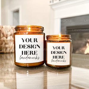 May include: Two lit amber glass jar candles with white labels that read "YOUR DESIGN HERE lavidamocks". The candles are on a reflective surface, with a woven basket and a fireplace in the background. The candles are different sizes.