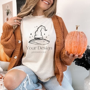 May include: A white t-shirt featuring a black and white illustration of a witch's hat and the text "Your Design" and "LAVIDAMOCKS". The model is wearing the t-shirt with a brown cardigan and blue jeans. A small orange pumpkin is being held in the model's hand.