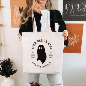 Peut inclure: Un sac fourre-tout blanc avec des anses noires et un motif de fantôme noir. Le sac porte l'inscription "Your Design Here" et "Est. 2024" imprimée.