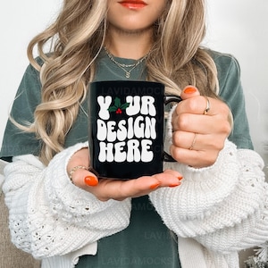 May include: A black ceramic mug with a white design that says "Your Design Here" with a small holly sprig. The mug is being held by a person wearing a green sweater and a white cardigan.