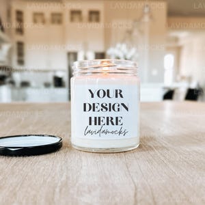 May include: A glass jar candle with a white label bearing the text "YOUR DESIGN HERE lavidamocks." The candle is on a wooden surface, with its black lid beside it. The background is blurred, suggesting a domestic setting.