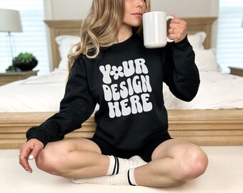 May include: A black sweatshirt with the text "YOUR DESIGN HERE" in white, retro-style lettering. The sweatshirt is being worn by a person sitting on a bed.