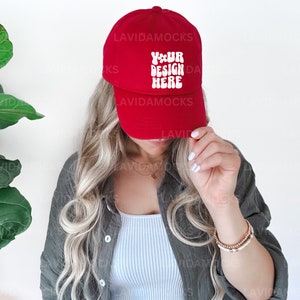 以下が含まれることがあります： 白い文字で「YOUR DESIGN HERE」と書かれた赤いベースボールキャップ。長いブロンドの髪をした人がキャップを持っています。