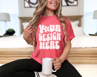 May include: A woman wearing a coral pink t-shirt with the text "YOUR DESIGN HERE" in white. She is holding a white mug and sitting on a bed.