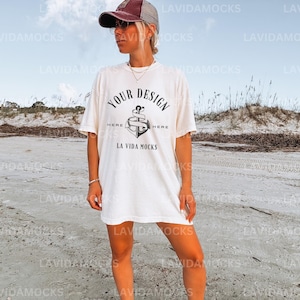 May include: A white oversized t-shirt with a graphic of an anchor and the text "YOUR DESIGN HERE HERE LA VIDA MOCKS". The model is wearing a brown baseball cap and sunglasses.