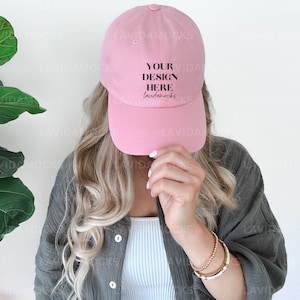 May include: A pink baseball cap with the text "YOUR DESIGN HERE" in black lettering. The cap is being held by a person with long blonde hair.