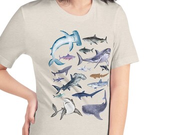 May include: A light beige t-shirt with a graphic print of various types of sharks in shades of blue, gray, and white. The sharks are arranged in a square pattern on the front of the shirt.