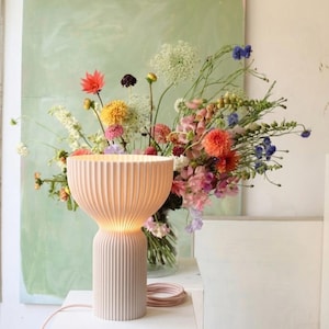 Könnte beinhalten: Eine hellrosa Tischlampe mit geriffeltem Schirm und Sockel, von innen beleuchtet. Ein bunter Blumenstrauß steht im Hintergrund. Die Lampe steht auf einer weißen Oberfläche mit einem rosa Kabel.