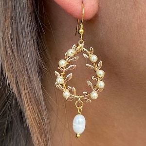 Peut inclure: Boucles d'oreilles dorées avec un motif de couronne, ornées de détails de feuilles et de petites perles rondes. Une seule perle en forme de goutte pend en bas. Les boucles d'oreilles sont présentées sur un fond flou.