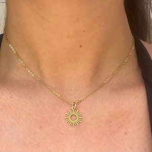 May include: A gold-colored necklace with a sunburst pendant. The pendant has a circular design with radiating lines and small beaded accents. The necklace is worn on a person's neck, against a black top.