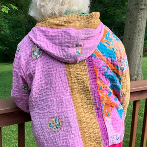 Puede incluir: Chaqueta acolchada con capucha, con un dise&ntilde;o de patchwork en tonos rosa, dorado, naranja y azul. La chaqueta tiene un aspecto artesanal &uacute;nico con costuras visibles y detalles bordados circulares. La prenda se lleva sobre pantalones rosas.