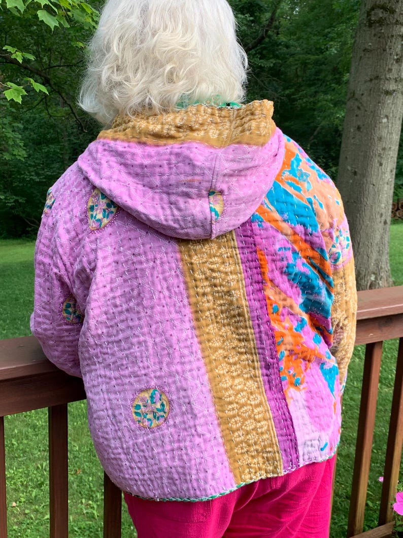 Puede incluir: Chaqueta acolchada con capucha, con un dise&ntilde;o de patchwork en tonos rosa, dorado, naranja y azul. La chaqueta tiene un aspecto artesanal &uacute;nico con costuras visibles y detalles bordados circulares. La prenda se lleva sobre pantalones rosas.