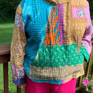 Puede incluir: Una sudadera con capucha de patchwork en tonos de azul, verde, morado y dorado. La sudadera con capucha tiene un bolsillo de canguro y una capucha con cord&oacute;n. La tela parece acolchada con costuras visibles. Las mangas y el cuerpo est&aacute;n hechos de diferentes telas estampadas.