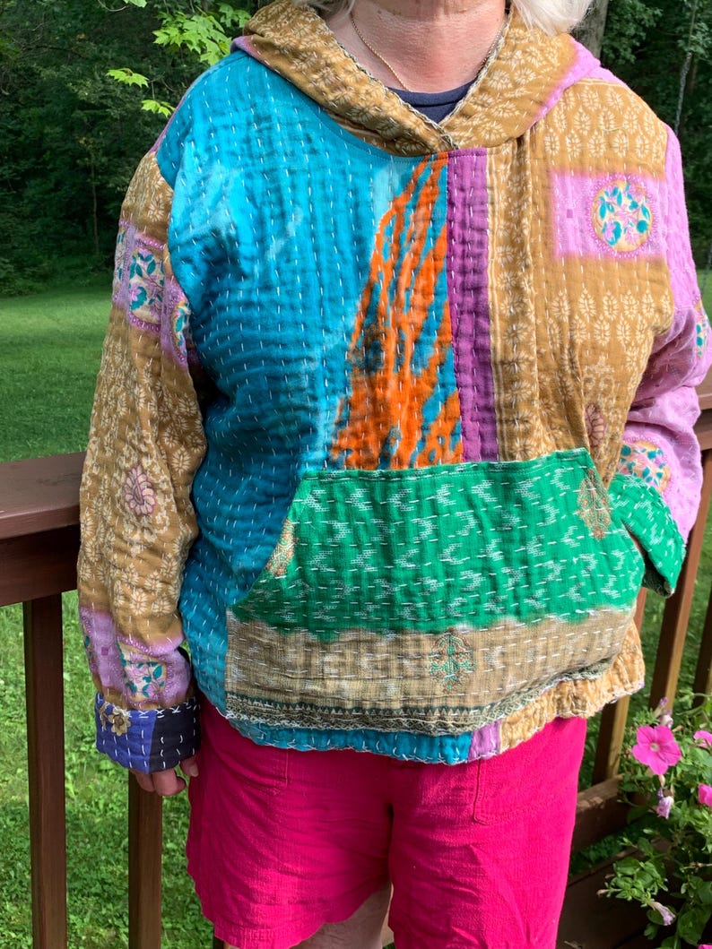 Puede incluir: Una sudadera con capucha de patchwork en tonos de azul, verde, morado y dorado. La sudadera con capucha tiene un bolsillo de canguro y una capucha con cord&oacute;n. La tela parece acolchada con costuras visibles. Las mangas y el cuerpo est&aacute;n hechos de diferentes telas estampadas.