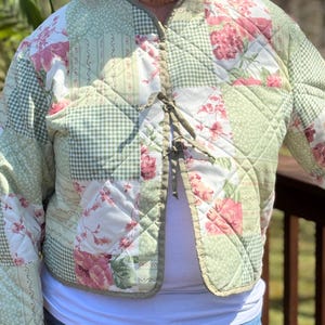 May include: A quilted jacket with a patchwork design. The jacket features a mix of floral, gingham, and striped patterns in shades of green, pink, and white. It has a tie closure at the front and a quilted texture.