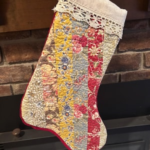 Puede incluir: Calcetín navideño acolchado con diseño patchwork. El calcetín presenta rayas verticales de tela floral en rojo, amarillo, azul y marrón. La parte superior es de color lino natural con un ribete de encaje y un borde rojo.