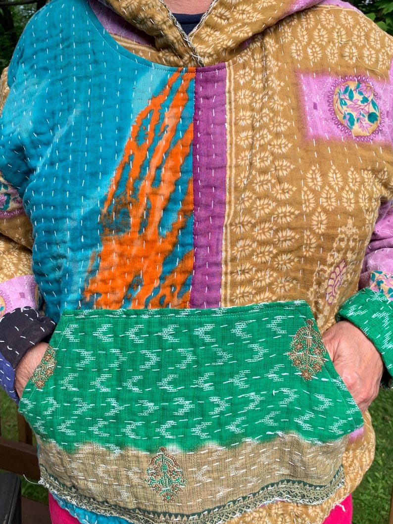 Puede incluir: Una sudadera con capucha de patchwork con bolsillo de canguro. La sudadera presenta una mezcla de colores y patrones, incluyendo turquesa, naranja, morado y dorado. La tela parece acolchada, con costuras visibles. El bolsillo es verde con un patr&oacute;n blanco.