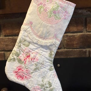 Puede incluir: Un calcetín navideño acolchado con un estampado floral. La sección superior presenta una cesta de flores rosa bordada. El calcetín es blanco con rosas rosas y verdes y un ribete de encaje. Un patrón de cuadros rosa y blanco bordea la parte superior.