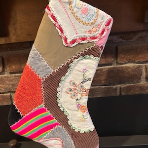 May include: A handmade Christmas stocking with a patchwork design. The stocking features various fabrics, including a floral embroidered section, a red textured section, and a pink and green striped section. The top is trimmed with lace and a floral pattern.