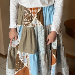 May include: A patchwork skirt featuring various fabric patterns and colors, including brown, blue, and floral prints. The skirt has a tiered design with a white eyelet lace trim at the hem. The skirt is paired with a white long-sleeved shirt.