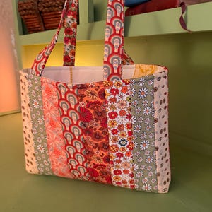 May include: A patchwork tote bag with a variety of floral and geometric patterns in shades of orange, red, green, and cream. The bag has two handles with a matching floral design. The interior is white.