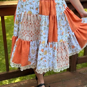 May include: A tiered midi skirt featuring a patchwork design of solid orange fabric, white fabric with an orange and yellow floral print, and cream-colored crochet lace panels. The skirt has an elastic waistband and a scalloped crochet lace hem.