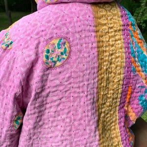 Puede incluir: Chaqueta acolchada en tonos rosa, dorado, morado y turquesa. La chaqueta presenta medallones florales bordados y un patr&oacute;n geom&eacute;trico cosido. La capucha y las mangas son visibles, mostrando el dise&ntilde;o patchwork y las texturas.