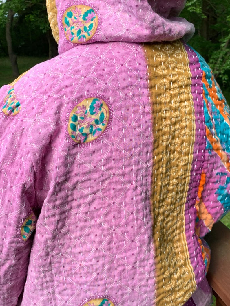 Puede incluir: Chaqueta acolchada en tonos rosa, dorado, morado y turquesa. La chaqueta presenta medallones florales bordados y un patr&oacute;n geom&eacute;trico cosido. La capucha y las mangas son visibles, mostrando el dise&ntilde;o patchwork y las texturas.