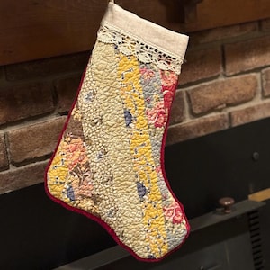 Può includere: Una calza di Natale trapuntata con un design patchwork. La calza presenta una parte superiore color crema con finiture in pizzo e un bordo rosso. Il patchwork include tessuti gialli, floreali e fantasia.
