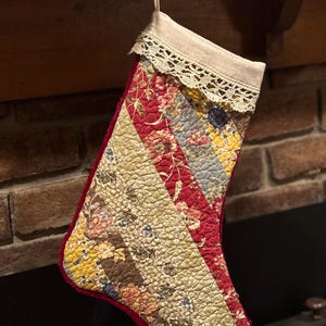 Könnte beinhalten: Ein gesteckter Weihnachtsstrumpf mit Patchwork-Design. Der Strumpf hat verschiedene Blumenmuster in Rot, Gelb und Beige mit rotem Rand. Die Oberseite ist aus Naturleinen mit Spitzenbesatz und einer Schlaufe zum Aufhängen.