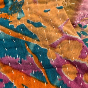 Puede incluir: Primer plano de un textil colorido con un dise&ntilde;o de patchwork. La tela presenta una vibrante mezcla de tonos turquesa, naranja y morado, con costuras blancas. El textil tiene un aspecto suave y texturizado.