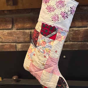 May include: Handmade Christmas stocking with a patchwork design. The stocking has a white top with pink floral lace trim. The body is made of various fabric patches in floral, pink, and red patterns.