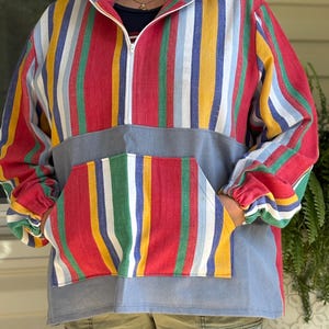 Puede incluir: Un jersey de rayas de colores con cierre de cremallera y un gran bolsillo en la parte delantera. El jersey está hecho de una tela suave y cómoda y tiene un corte holgado.