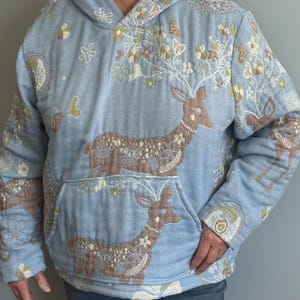 May include: A light blue hooded sweatshirt with a floral and deer design. The deer are embroidered in brown and white with floral accents. The sweatshirt has a kangaroo pocket.