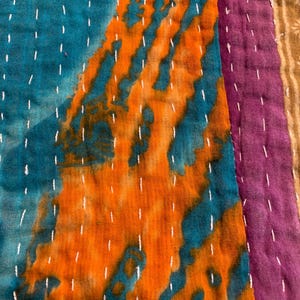 Puede incluir: Primer plano de un textil patchwork con colores vibrantes. El dise&ntilde;o presenta secciones de tela turquesa, naranja, morada y verde. Las costuras blancas acent&uacute;an las costuras y los patrones. El textil tiene una apariencia texturizada, lo que sugiere una calidad artesanal.
