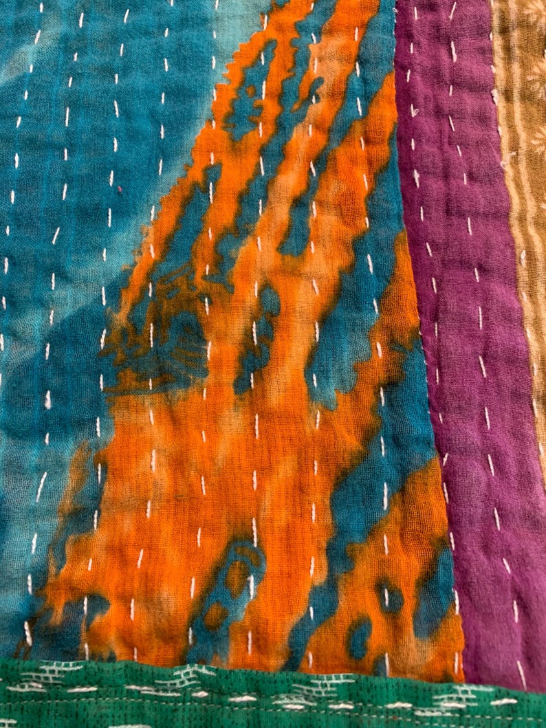 Puede incluir: Primer plano de un textil patchwork con colores vibrantes. El dise&ntilde;o presenta secciones de tela turquesa, naranja, morada y verde. Las costuras blancas acent&uacute;an las costuras y los patrones. El textil tiene una apariencia texturizada, lo que sugiere una calidad artesanal.
