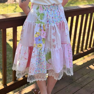 Op de afbeelding: Een patchwork rok met een gelaagd ontwerp, met verschillende bloemen- en patroonstoffen in roze, wit en groen. De rok heeft een kanten rand aan de zoom. De persoon draagt bruine sandalen.
