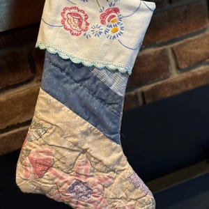 May include: A handmade Christmas stocking with a patchwork design. The top is white with embroidered flowers and a blue lace trim. The body features blue and cream quilted fabric with floral patterns.