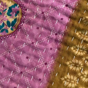 Puede incluir: Primer plano de una tela acolchada con un medall&oacute;n floral en oro, verde azulado y rosa. La tela presenta una combinaci&oacute;n de colores rosa y dorado con costuras blancas. El dise&ntilde;o incluye patrones geom&eacute;tricos y motivos florales.