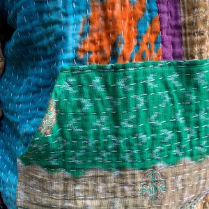 Puede incluir: Una chaqueta patchwork con una vibrante mezcla de colores y texturas. La chaqueta presenta un patr&oacute;n te&ntilde;ido anudado en azul turquesa y naranja, un bolsillo verde y secciones a rayas marrones y moradas. Costuras blancas detallan las costuras.